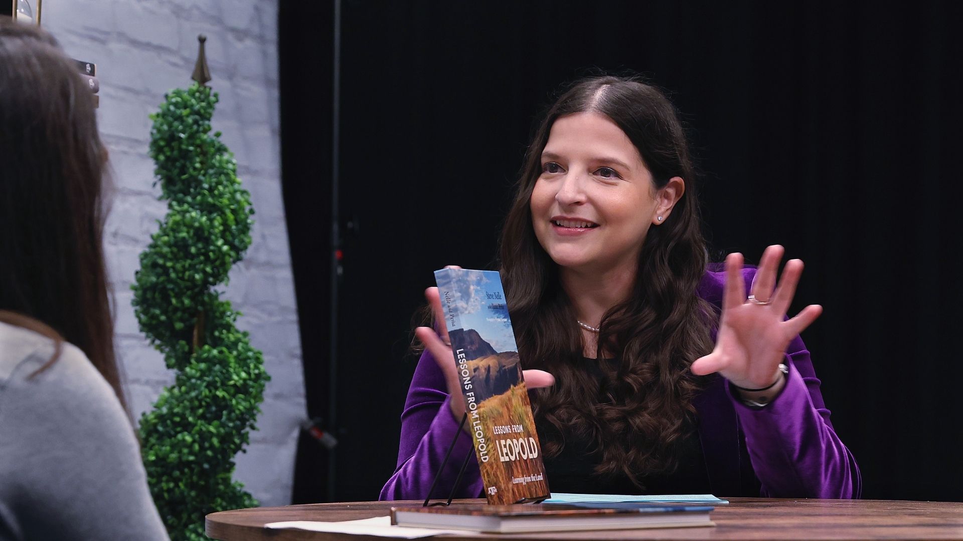 Woman on a recording studio stage sits at a table, facing another speaker. A book, "Lessons From Leopold," is on a display stand between them.
